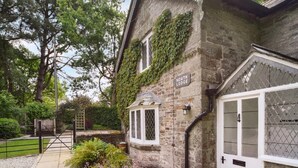 A stone building with a sign reading 'North Lodge,' a gated entrance, and a small garden with pink flowers. - 2 Bed in Launceston (oc-84660) (Tregeare)