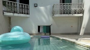 Uma piscina com piso ladrilhado, um brinquedo flutuante azul insuflável e um prédio com varandas.