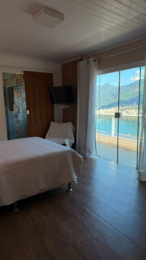 Habitación de hotel con una cama, televisor montado en la pared y un ventanal con vista a un cuerpo de agua y montañas.