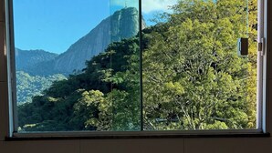 Quarto com uma janela grande que oferece vista para uma montanha e vegetação exuberante.
