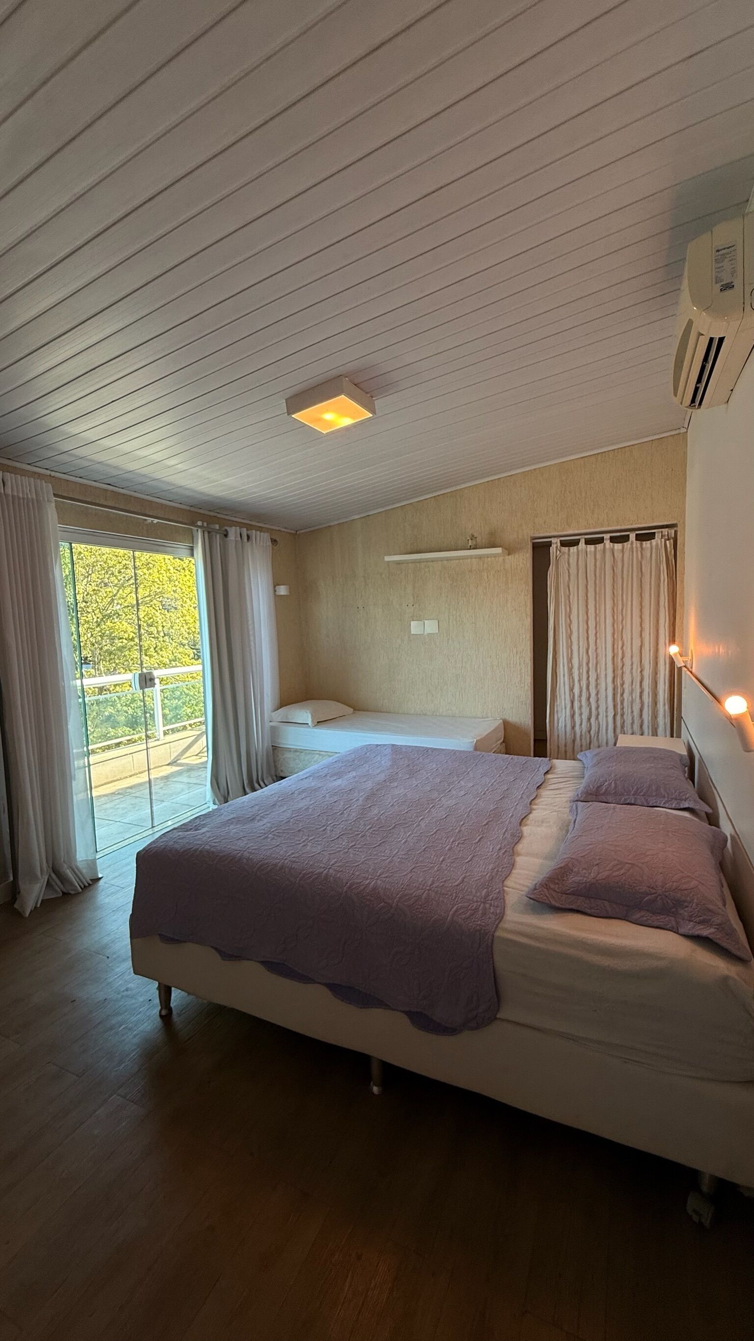 A bedroom with a large bed, a smaller bed, a ceiling fan, an air conditioning unit, and a view of the outdoors through a sliding door.