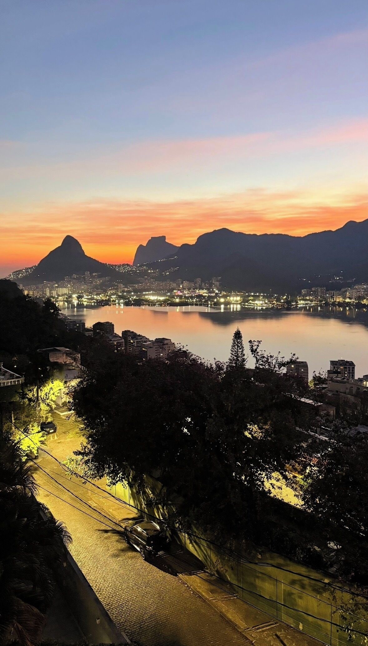 A cityscape at dusk with a prominent mountain silhouette, a calm river reflecting city lights, and a street with a tram.