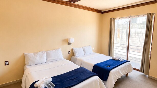 A hotel room with two beds, a ceiling vent, a window with curtains, and a wall-mounted light.