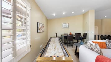 A room with a shuffleboard table, a dining table with chairs, and a sofa with pillows.