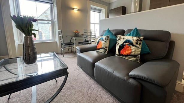 Ein Wohnzimmer mit Glastisch, einer schwarzen Ledersofa mit dekorativen Kissen, einem Essbereich mit Tisch und Stühlen sowie einem Fenster mit Blick.