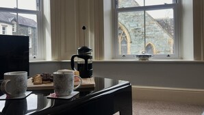 Una habitación con piano, cafetera y vista a un edificio de piedra a través de amplios ventanales.
