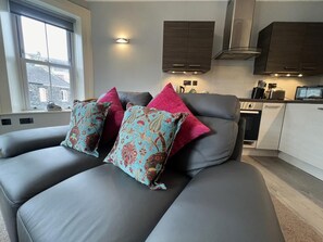 A modern living room with a grey sofa, patterned cushions, a kitchen area with cabinets and appliances, and a window with a view. - 1 Bed in Keswick (oc-sz157) (Keswick)