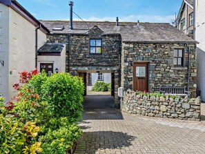 A stone building with a central entrance, a chimney, and a small garden area with plants and flowers. - 2 Bed in Keswick (oc-sz449) (Keswick)