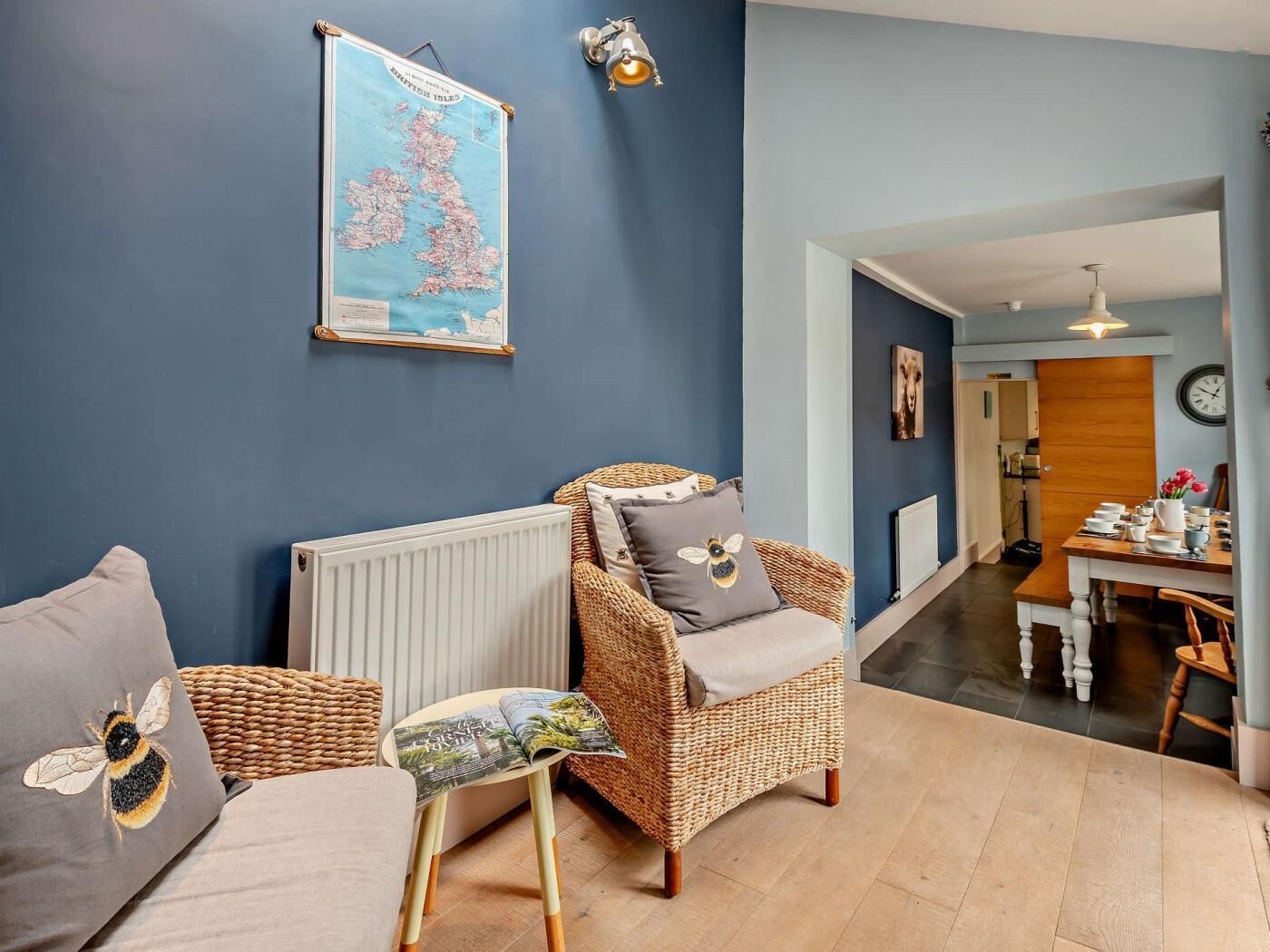Un salon avec un mur bleu, une carte, un fauteuil en osier, un canapé et, au fond, un coin repas.