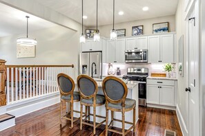 A kitchen with white cabinets, a stainless steel refrigerator, a microwave, and a stove. There are three wooden chairs with grey cushions. - The Clifton | Bars, Bourbon, and Brunch on Foot | Sleeps 15 (Louisville)