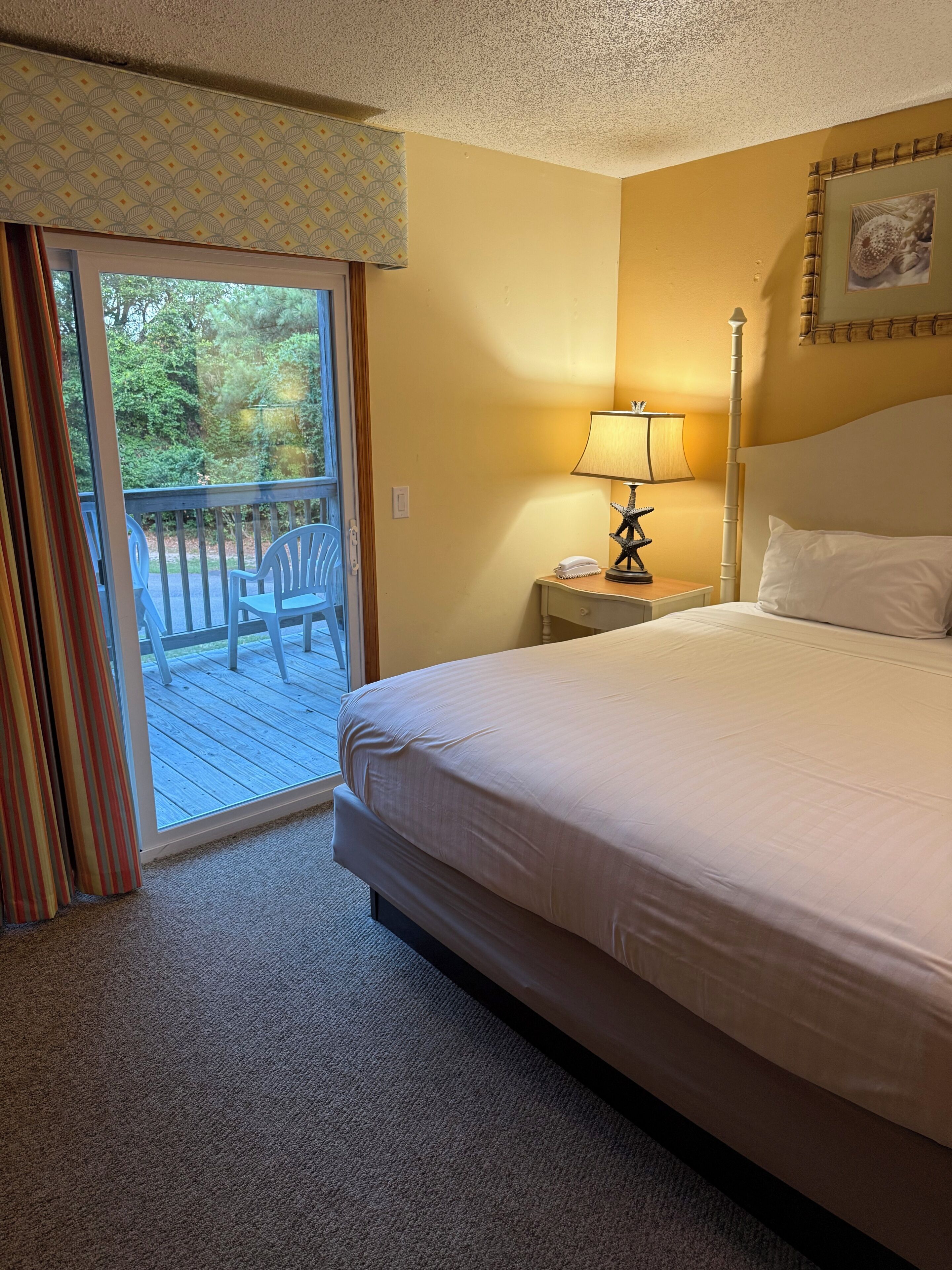 A hotel room with a bed, a nightstand, a lamp, a picture on the wall, and a balcony with chairs.