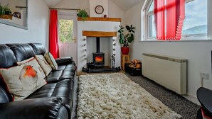 A cozy living room with a fireplace, a leather sofa, a fluffy rug, and a large window with red curtains. - 1 Bed in Penrith (oc-sz254) (Hutton)