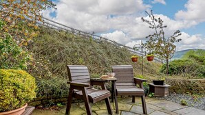 An outdoor patio with two wooden chairs and a small table, surrounded by stone flooring and potted plants. - 1 Bed in Penrith (oc-sz254) (Hutton)