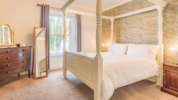 Ein Himmelbett mit Kopfteil, ein Schminktisch, ein Spiegel und ein Fenster mit Vorhängen.