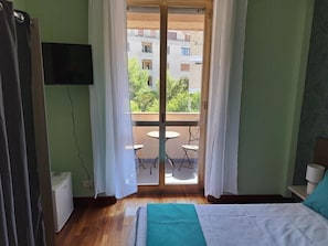 A bedroom with a bed, a window with curtains, a view of a balcony with outdoor furniture, and a television mounted on the wall. - B&B verdi (Lecce)