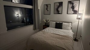 Une chambre avec une grande fenêtre, un lit recouvert de linge de lit blanc, une lampe sur pied et des œuvres d’art abstraites accrochées au mur.