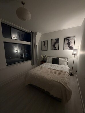 Ein Schlafzimmer mit großem Fenster, einem Bett mit weißer Bettwäsche, einer Stehlampe und abstrakten Wandbildern.