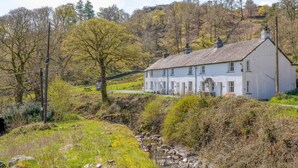 A white house with a gabled roof, surrounded by trees and a stream. - 3 Bed in Keswick (oc-sz234) (Borrowdale)