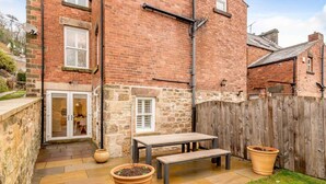 A brick house with a stone wall, a wooden fence, a patio with a table and two planters, and a door leading to a room. - 5 Bed in Matlock Bath (oc-85188) (Matlock Bath)