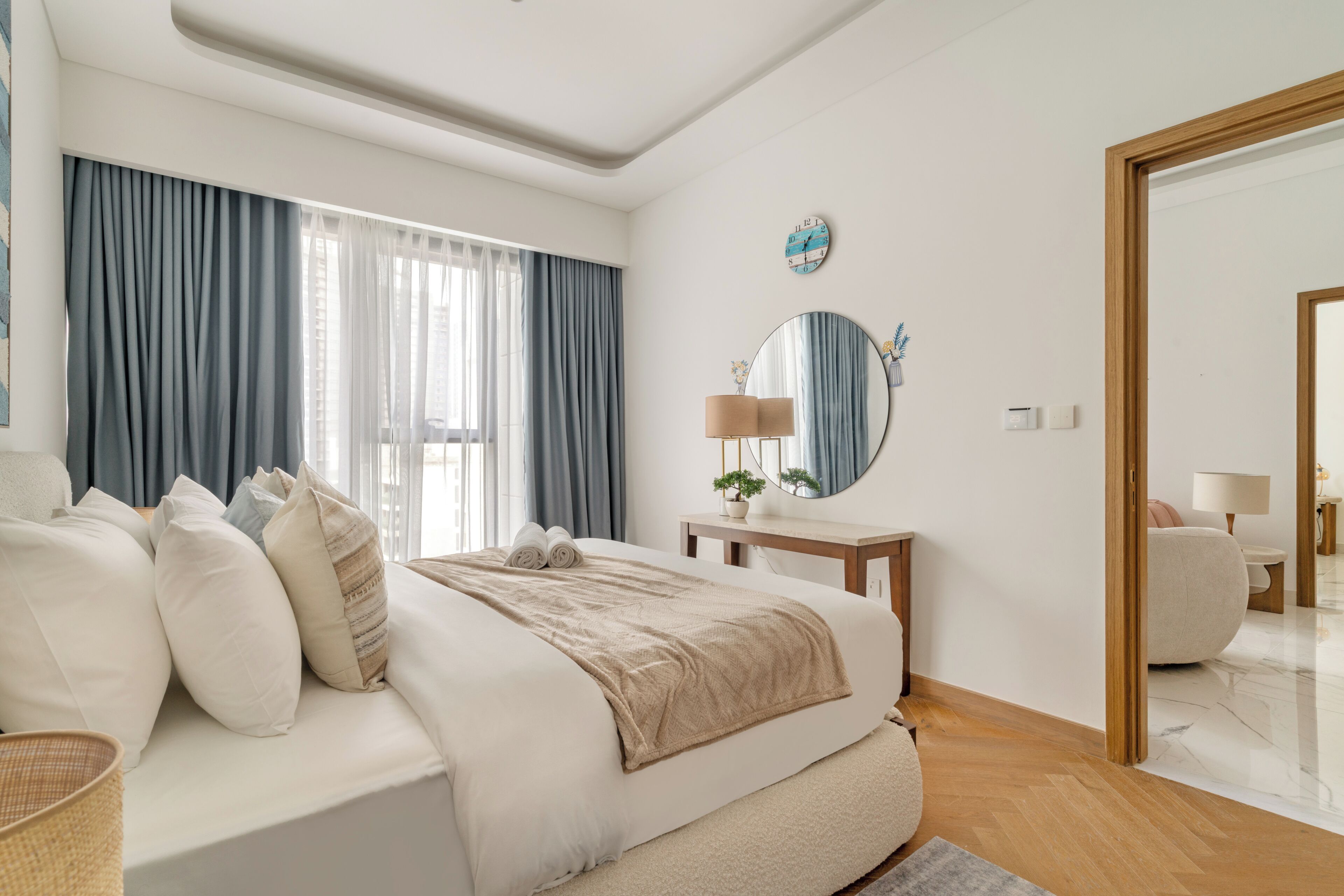 Une chambre moderne avec un grand lit, une table de chevet avec une lampe, un miroir et une vue sur une autre pièce par une porte ouverte.