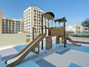 Ein Spielplatz mit Rutsche und Klettergerüsten, vor einer Kulisse aus modernen Gebäuden und blauem Himmel.