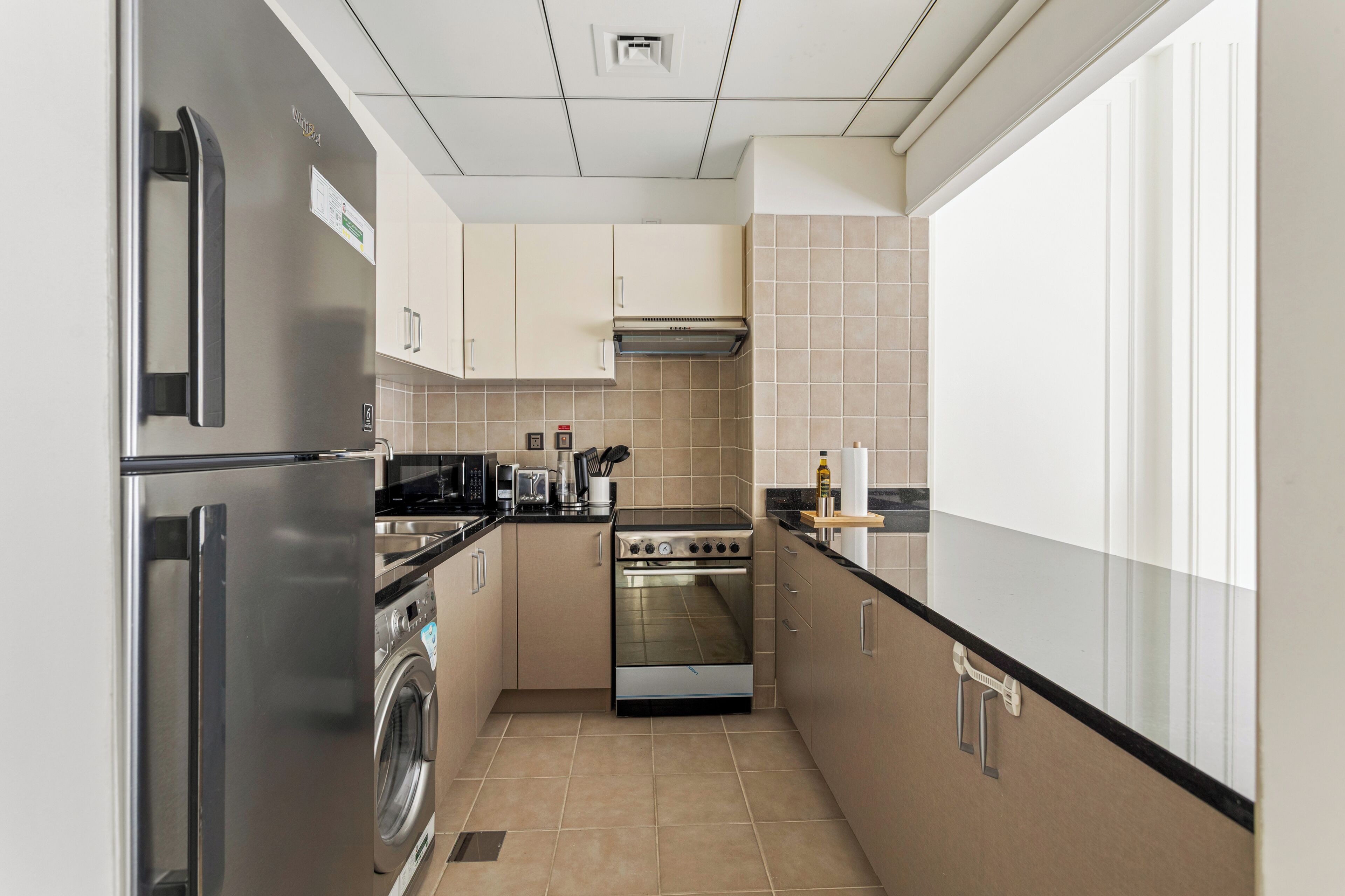 A compact kitchen with a stainless steel refrigerator, a built-in oven, a washing machine, and a microwave.