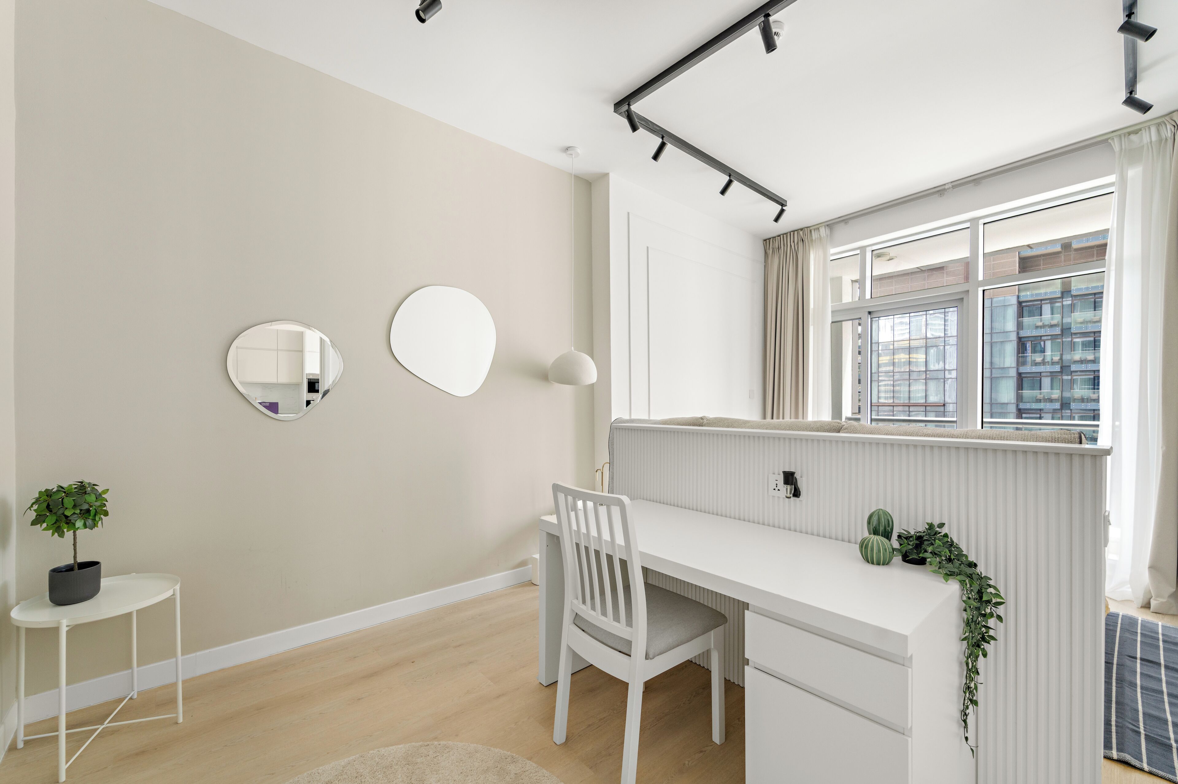 A minimalist child's room with a white desk, two wall mirrors, a potted plant, and a window with curtains.