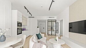 Uma sala de estar moderna com sofá, mesa de centro e um espaço de jantar com mesa e cadeiras. Há uma cozinha com forno e armários, e uma porta de vidro deslizante que dá para outro cômodo.