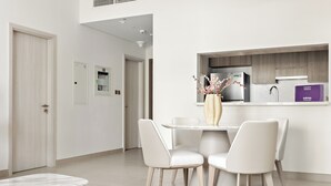 Un comedor moderno con una mesa redonda y cuatro sillas, un florero con flores, y una encimera de cocina con un refrigerador y una caja morada.