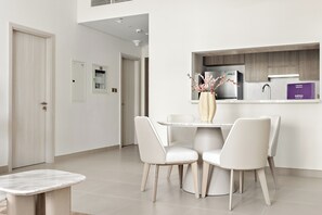 A modern dining area with a round table and four chairs, a vase with flowers, and a kitchen counter with a refrigerator and a purple box. - Silkhaus Yas Golf Collection (Abu Dhabi)