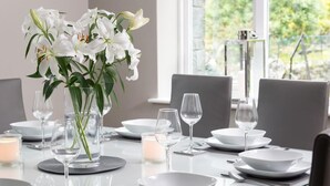 Una mesa de comedor dispuesta con platos, tazones y vasos blancos, un florero con flores blancas y una vela.