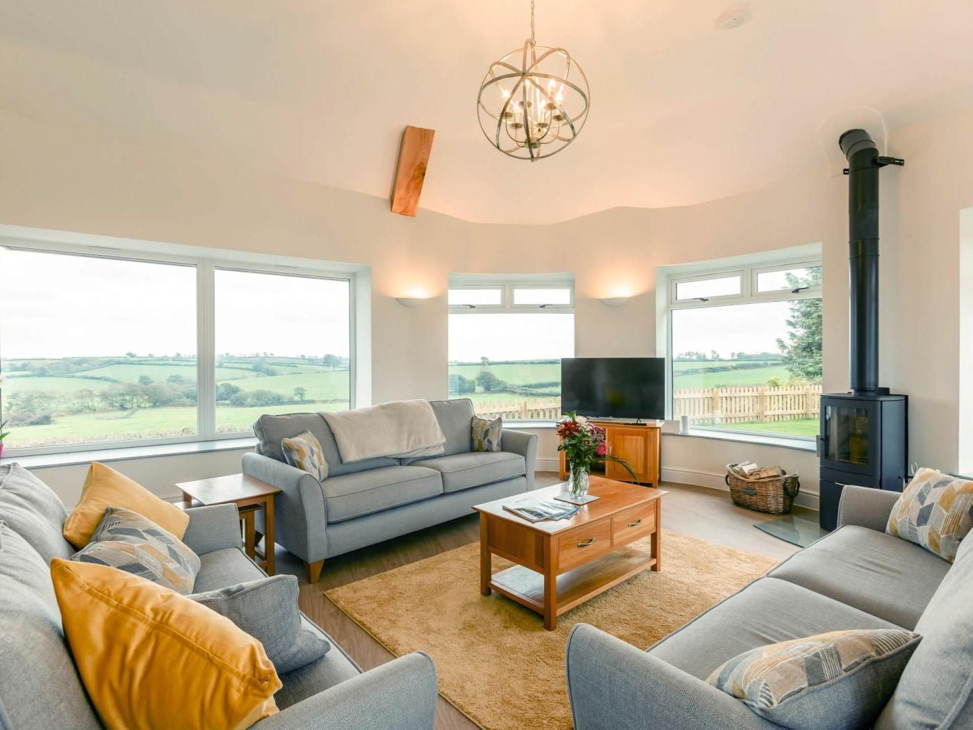 A modern living room with a sofa set, a television, a fireplace, and large windows offering a view of the countryside.