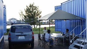 Un coche aparcado frente a un contenedor azul de transporte, una mesa bajo un toldo, y una persona sentada a la mesa.