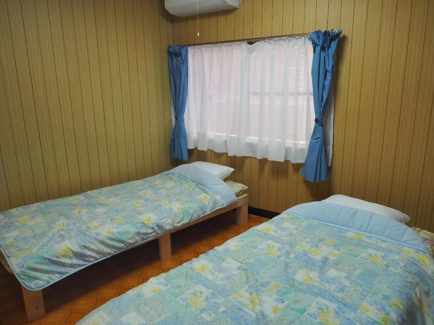 A room with two single beds, wooden flooring, a window with blue curtains, and a white air conditioning unit.