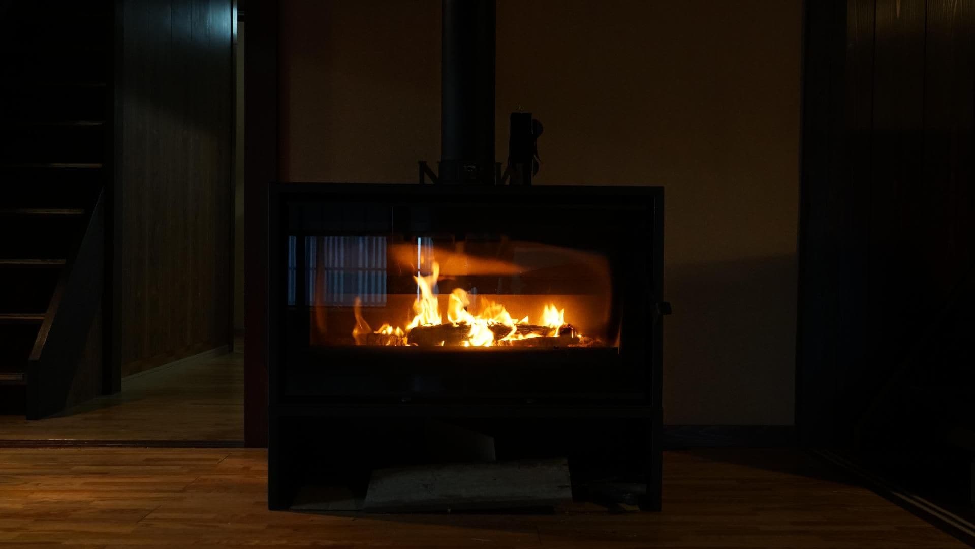 A wood-burning stove with visible flames, located in a room with wooden flooring and a staircase in the background.