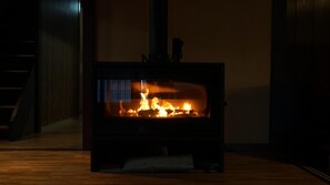 Ein Kaminofen mit sichtbaren Flammen, der sich in einem Raum mit Holzdielen befindet, im Hintergrund ist eine Treppe zu sehen.