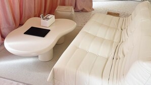 Un sofá blanco con un reposapiés, una tableta negra sobre el reposapiés, un baúl blanco y una cortina rosa de fondo.