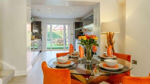 A modern dining area with a glass table, orange chairs, and a view into the kitchen and outdoor space. - 3 Bed in Porthcawl (oc-85193) (Drenewydd yn Notais)