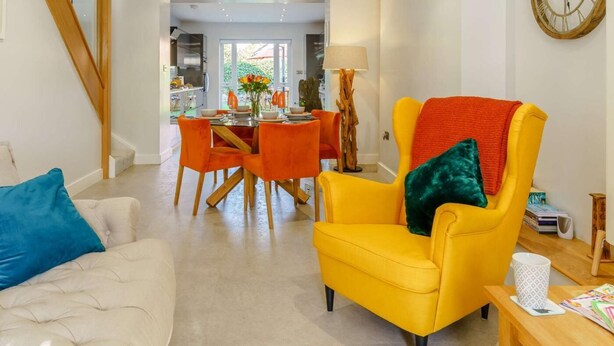 A modern living room with a yellow armchair, a white sofa, and a dining area with orange chairs. - 3 Bed in Porthcawl (oc-85193) (Drenewydd yn Notais)
