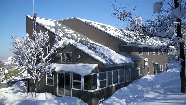 A two-story building with a sloped roof covered in snow, large windows, and a visible satellite dish. - Pension Mogu (Shinano)