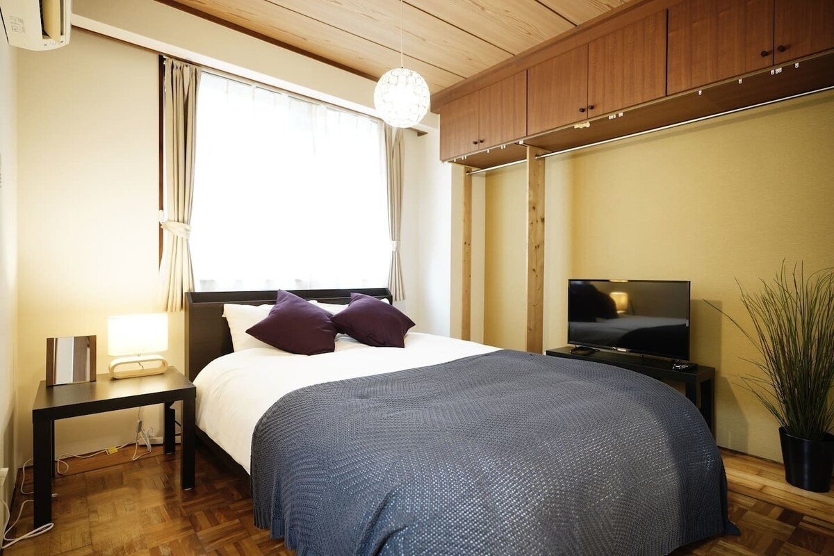 A bedroom with a bed, bedside table, lamp, TV, and a potted plant.