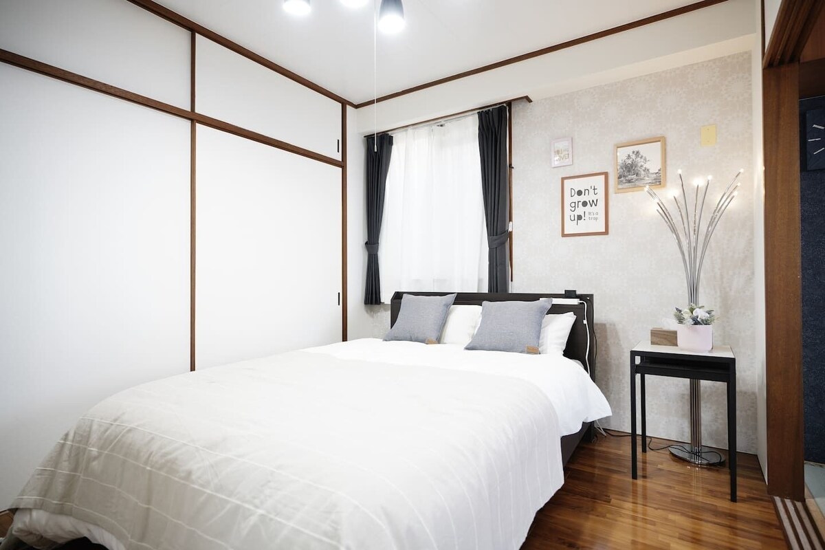 A bedroom with a bed, a bedside table, a window with curtains, and a wall with framed pictures.