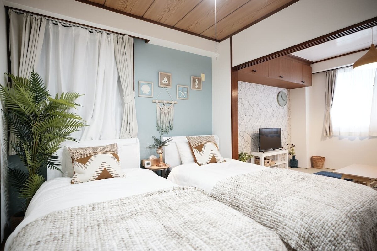 A bedroom with two beds, a TV, a desk, and a large plant.