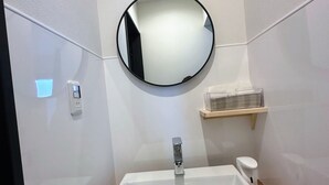 Un bany amb un lavabo blanc, un mirall rodó, una superfície de fusta, una petxina amb articles de tocador i una làmpada penjada a la paret.