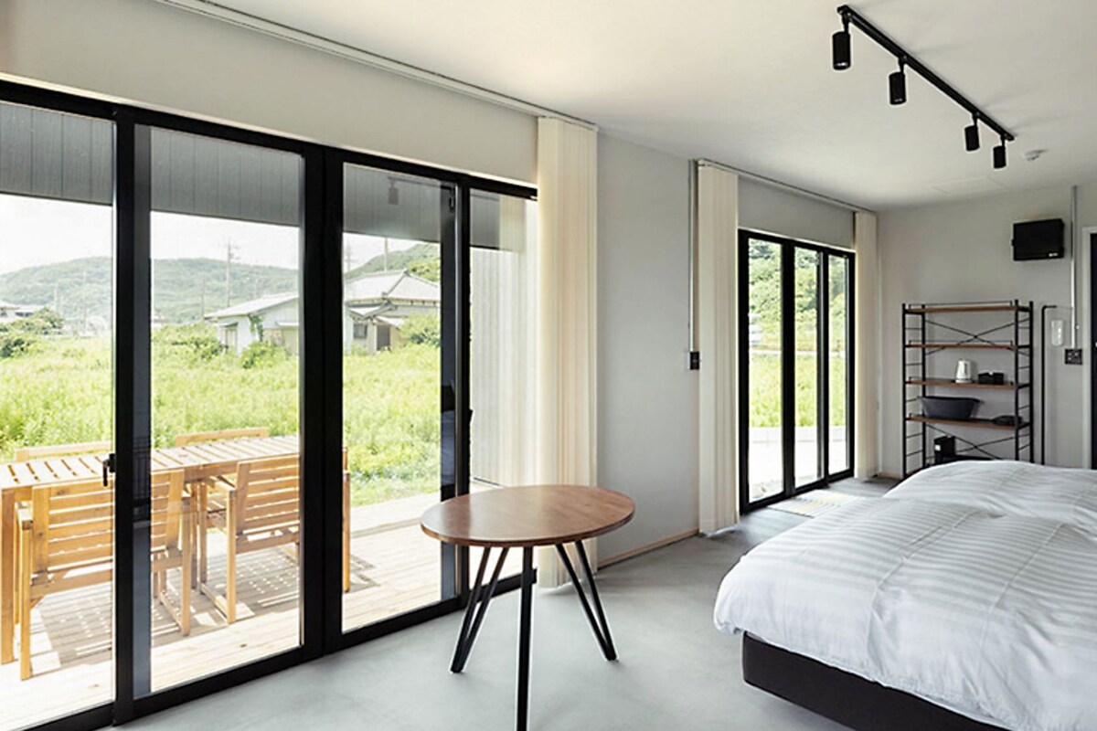 A modern bedroom with a large bed, a small round table, and a balcony with a view of greenery.