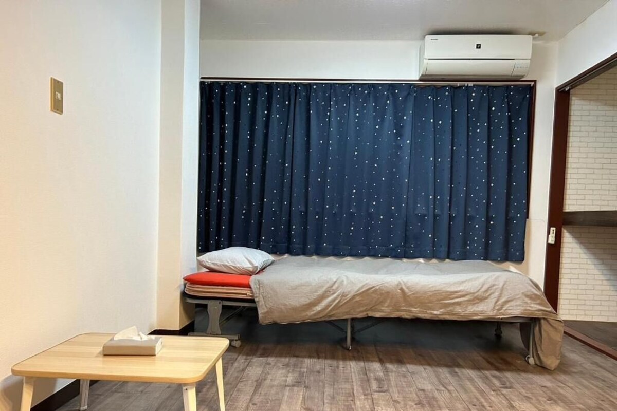 A small room with a bed, a table, a curtain with a star pattern, and an air conditioning unit.