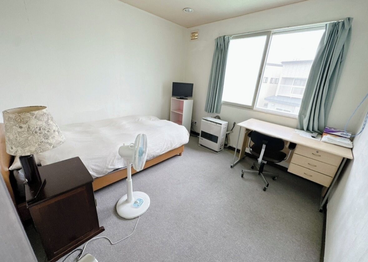 A hotel room with a bed, desk, chair, lamp, air conditioning unit, and a window with curtains.