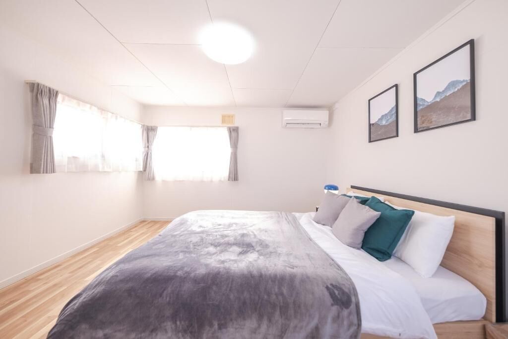 A modern bedroom with a wooden floor, a large bed with white and green pillows, a ceiling light, and two framed pictures on the wall.