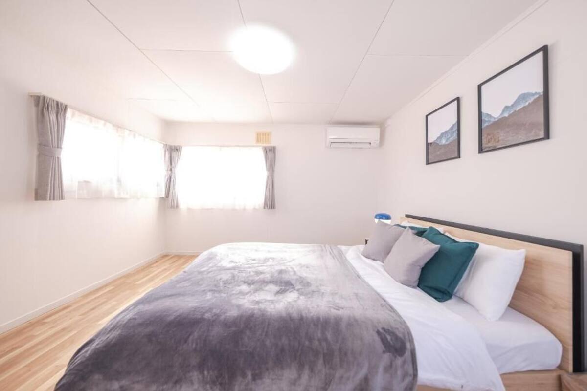Un dormitorio moderno con piso de madera, una cama grande con almohadas blancas y verdes, una lámpara de techo y dos cuadros en la pared.