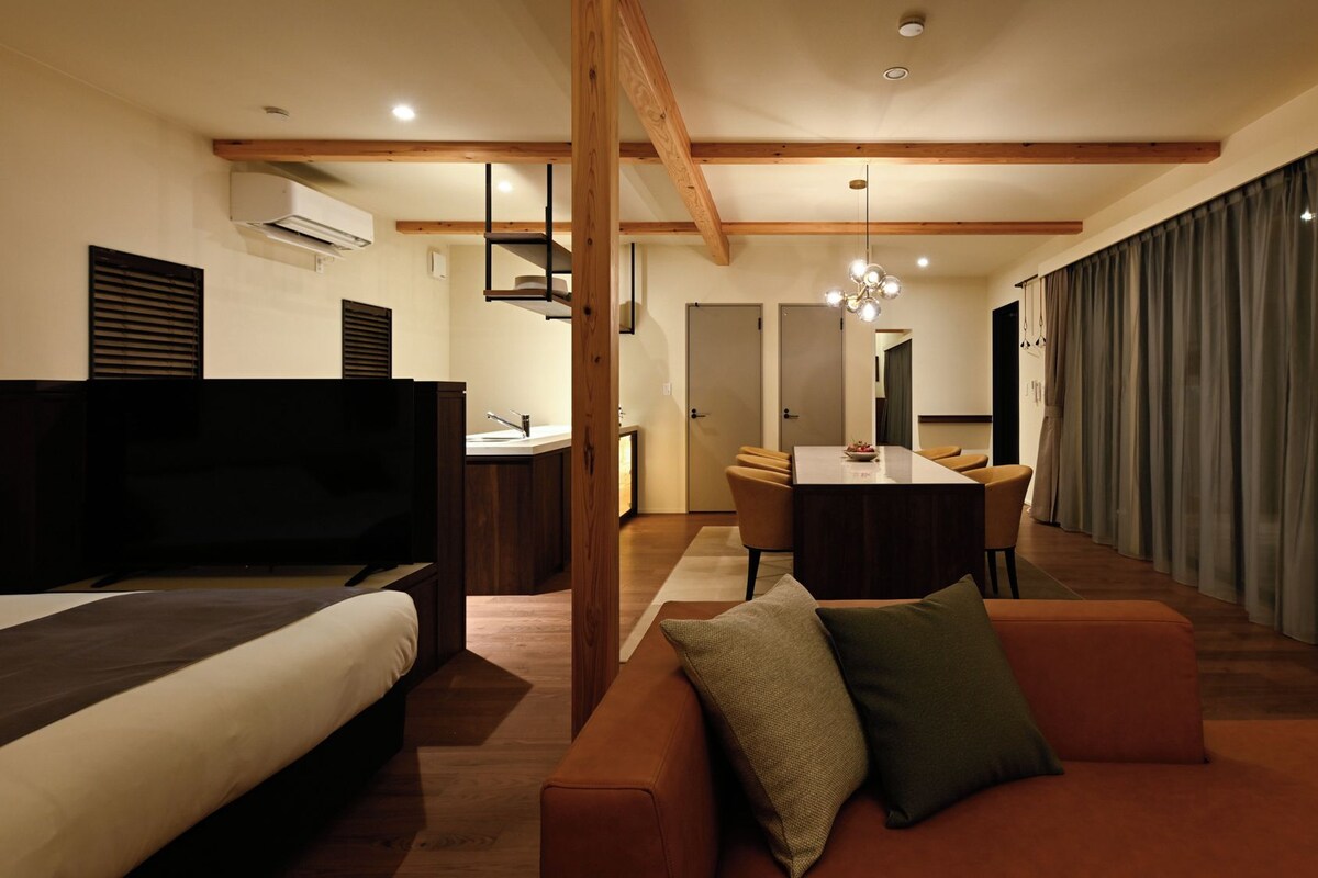 Ett modernt hotellrum med en säng, soffa, TV, matbord, stolar och ett stort fönster med gardiner.